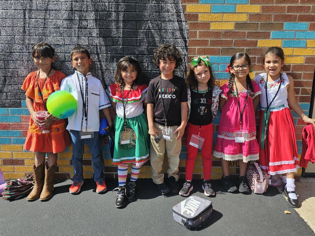 Seven smiling children pose outdoors in colorful cultural outfits beside a brick wall; one holds a green ball, with a lunch bag on the ground.