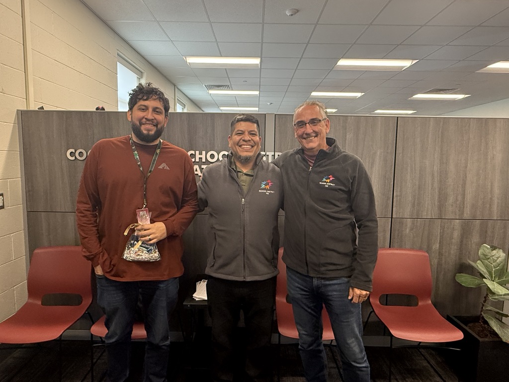 Three smiling men pose in an office hallway, one holding a small award, with red chairs and a plant behind them; friendly, celebratory mood.