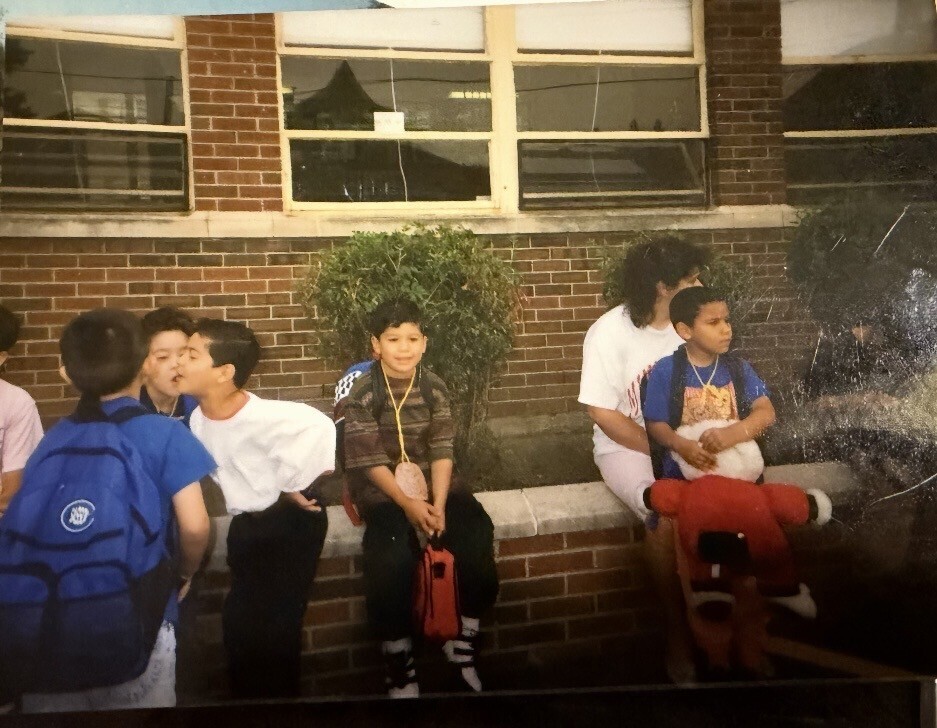 Children with backpacks sit and chat on a brick school ledge, while two students share a playful moment. Nostalgic, candid scene.