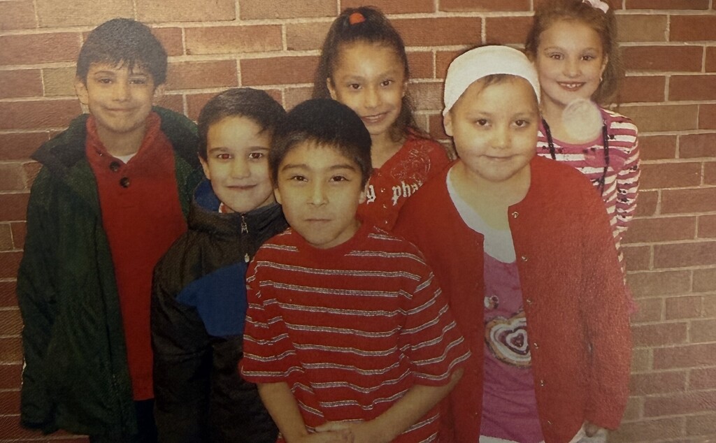 Six smiling children pose together against a brick wall, wearing colorful casual clothing in a warm, cheerful group portrait.