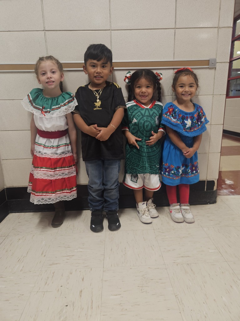 Four smiling children pose side by side in colorful traditional outfits, including embroidered dresses and a black shirt, in a school hallway.