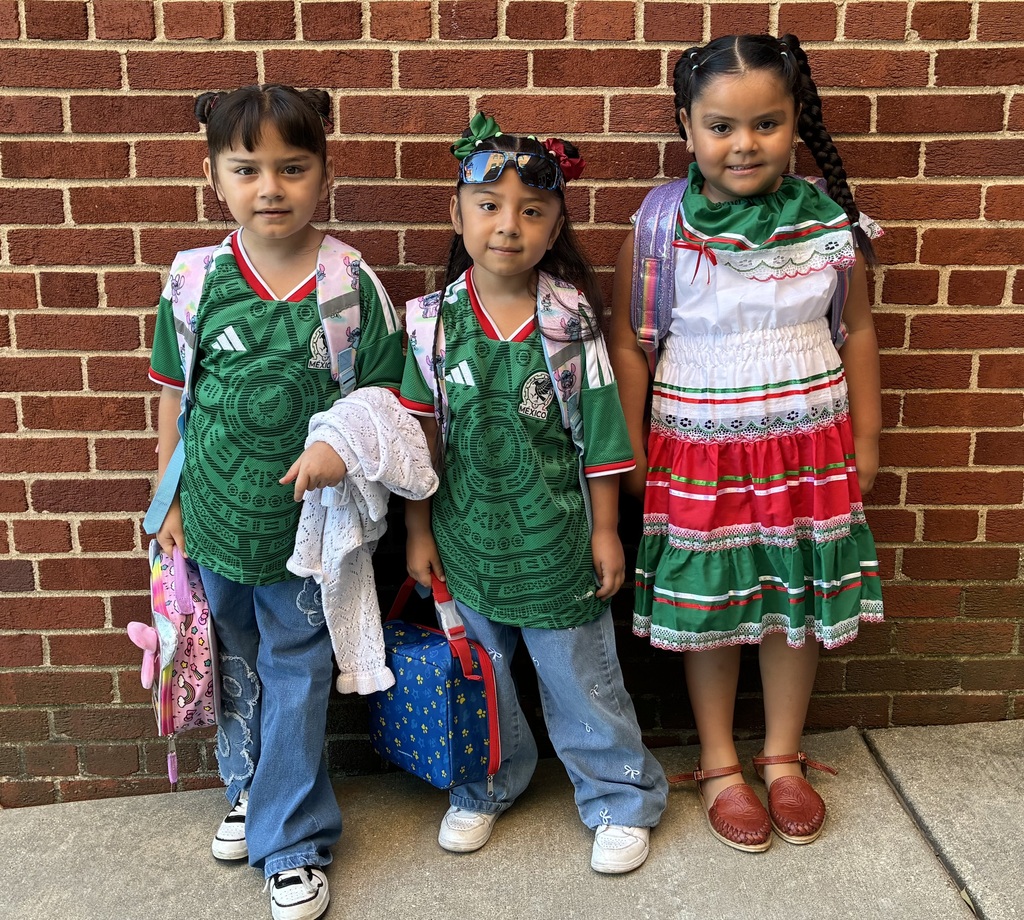 Three children pose cheerfully against a brick wall: two wear green soccer jerseys, while a girl wears a colorful traditional dress and red shoes.
