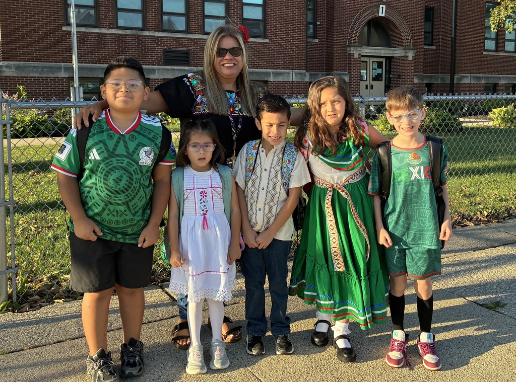 Six smiling children and an adult pose outside a brick school, wearing colorful cultural outfits on a sunny day; cheerful, celebratory mood.