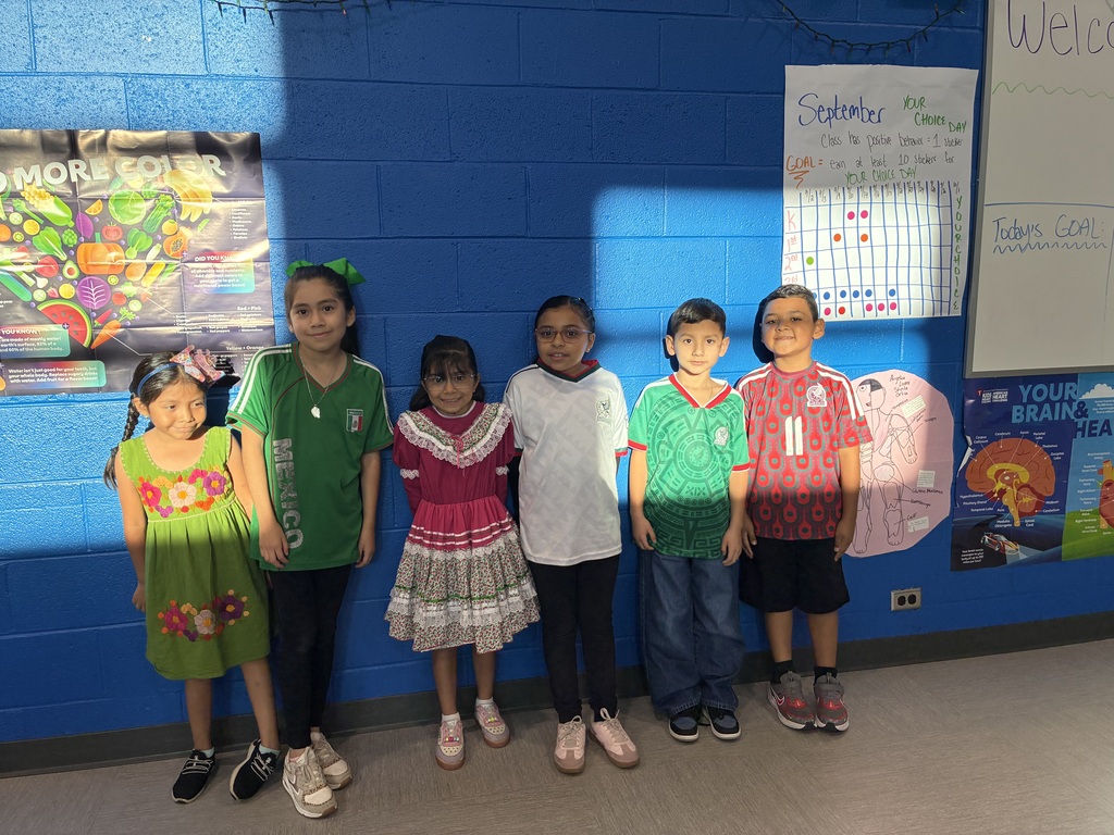 Six diverse children pose side by side in colorful clothing against a bright blue classroom wall, smiling beneath educational posters.