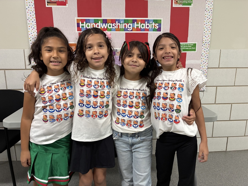 Four smiling schoolgirls pose arm-in-arm beneath a “Handwashing Habits” sign, wearing colorful hygiene-themed shirts in a classroom.