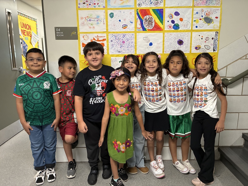 Eight smiling children pose in a school hallway beneath colorful student artwork, creating a cheerful, welcoming group portrait.