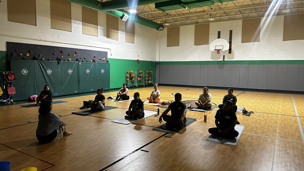 Children sit on yoga mats in a sunlit school gym, following an instructor during a calm mindfulness or stretching session.