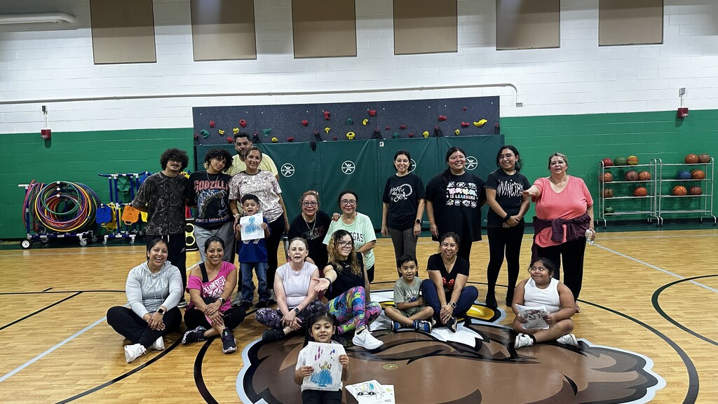 A diverse, cheerful group poses on a basketball court, with colorful climbing holds, sports equipment, and a bear logo on the floor behind them.