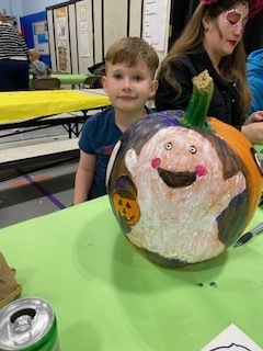 Young boy smiles beside a colorful painted pumpkin with a happy face and green stem on a table at a cheerful school craft event.