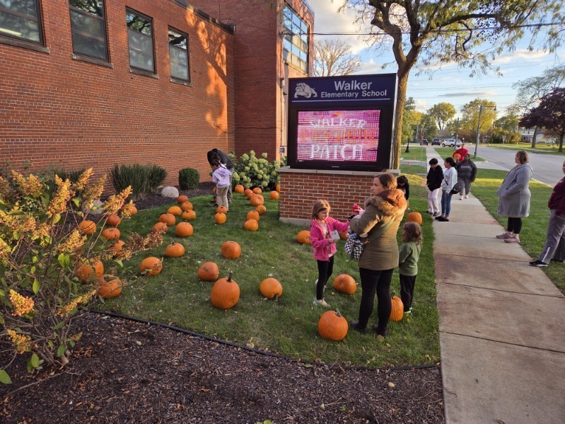Families gather outside Walker Elementary School among dozens of pumpkins, with children and adults enjoying a cheerful fall display.