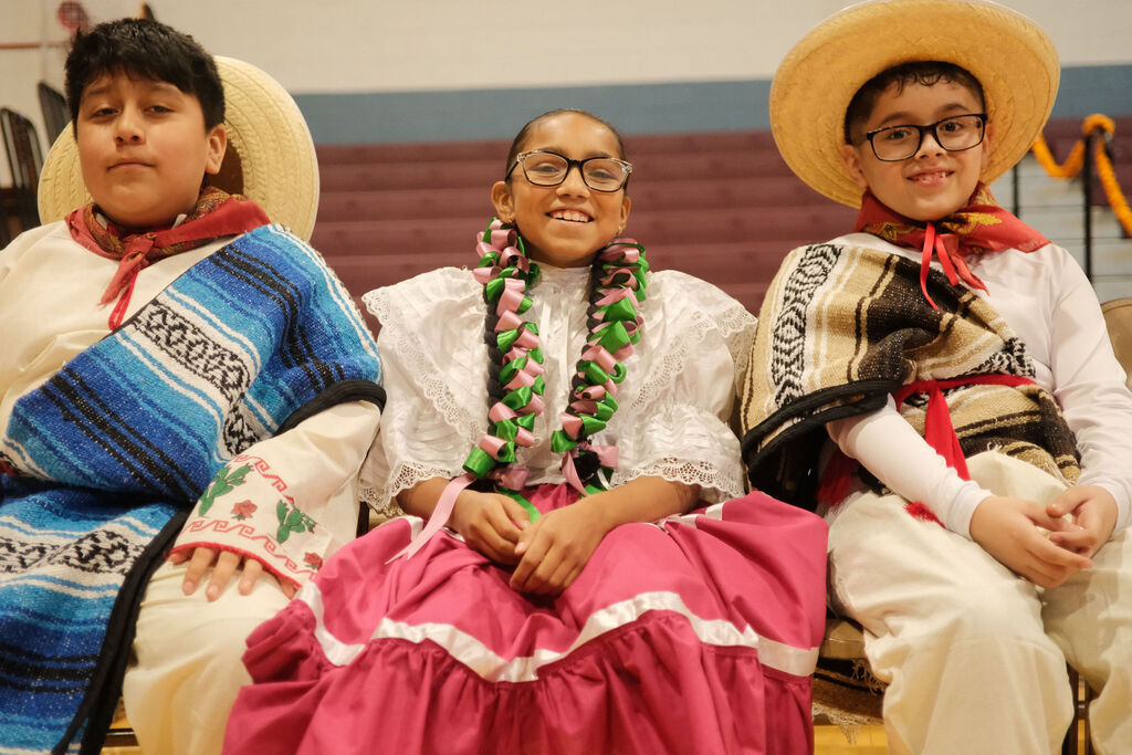 Three smiling children pose in colorful traditional Mexican clothing, sombreros, and floral leis, creating a festive, joyful cultural portrait.
