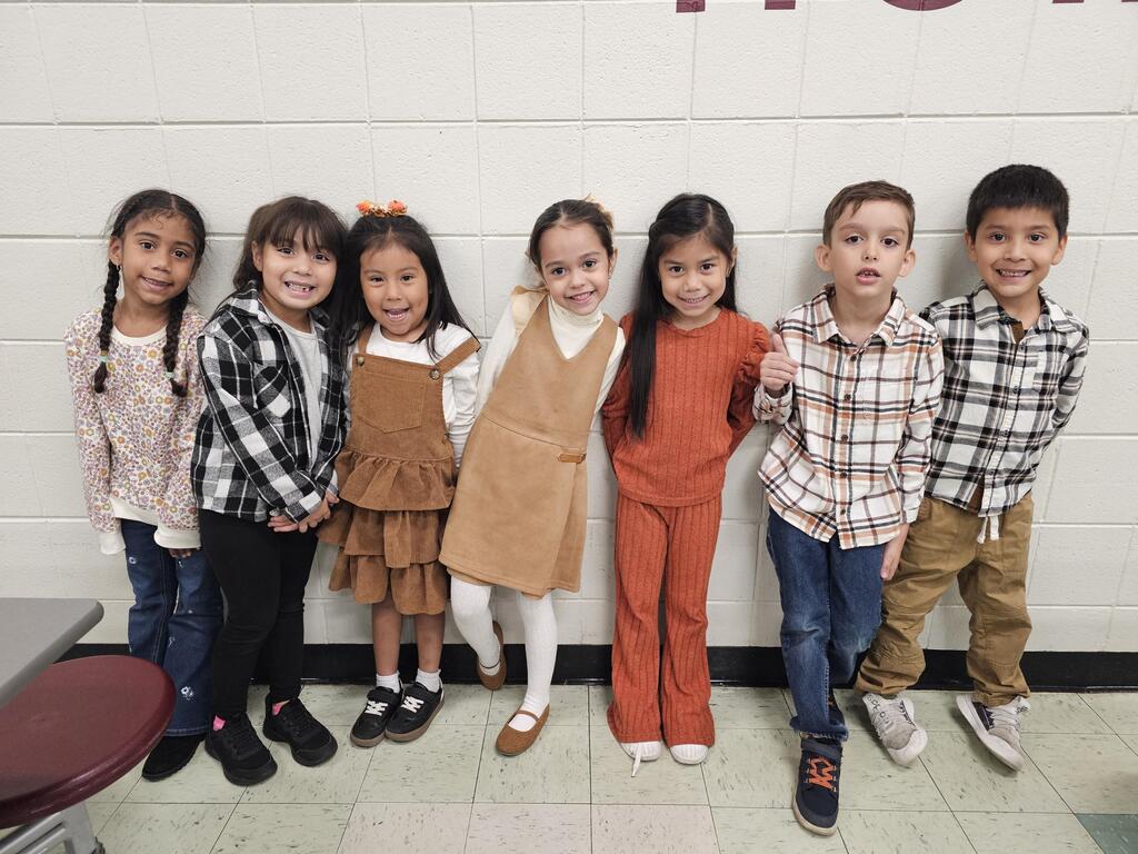 Seven smiling elementary-school children pose together indoors against a white brick wall, wearing colorful casual outfits, conveying joy and friendship.