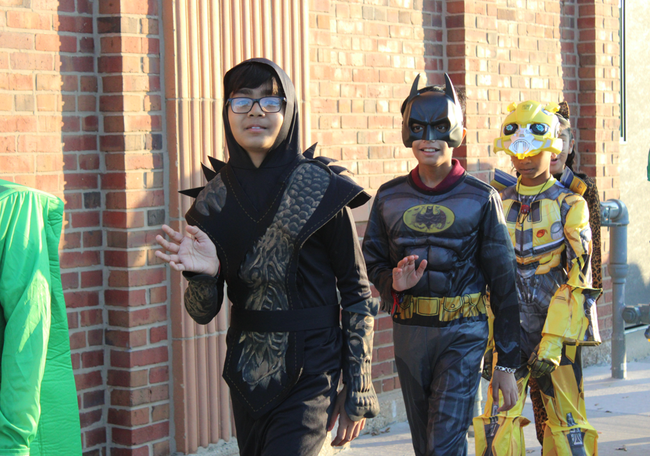 Three cosplayers walk beside a brick building: a black-armored figure, Batman, and a yellow robot, creating a playful mood.