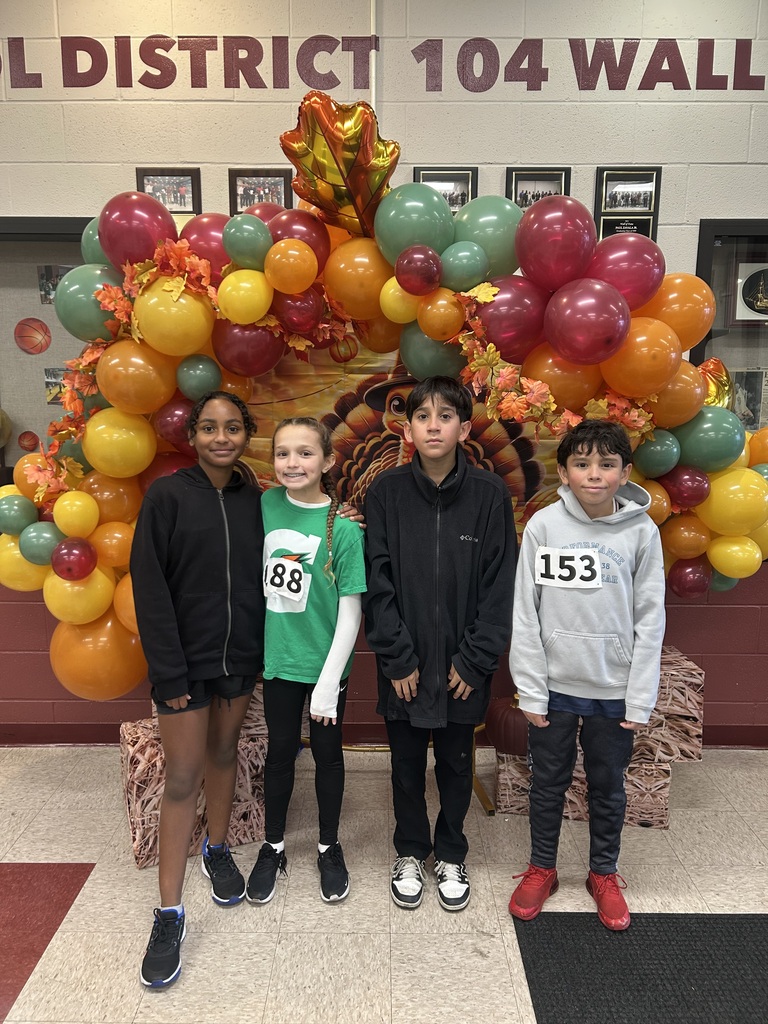 Four children smile beneath a colorful balloon arch, with fall decorations and race bibs, in front of a wall reading “District 104.”