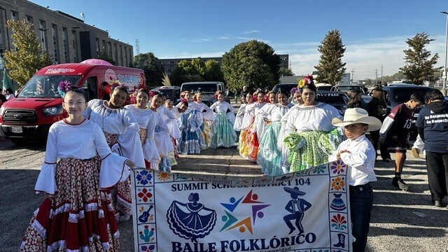 A cheerful folkloric dance group in colorful traditional costumes holds a “Baile Folklórico” banner in a sunny parking lot, ready to celebrate.