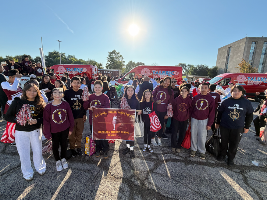 A large student group poses in burgundy and black school shirts beside a school banner and Taliercio vans, with bright sun overhead; upbeat community pride.
