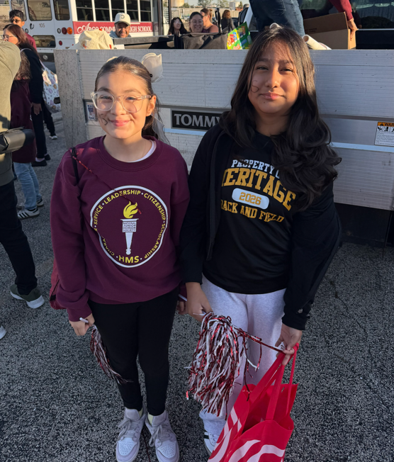 Two smiling students pose together at a busy outdoor school event, holding red-and-white pom-poms and a red bag; their casual school spirit shirts suggest a cheerful, festive mood.
