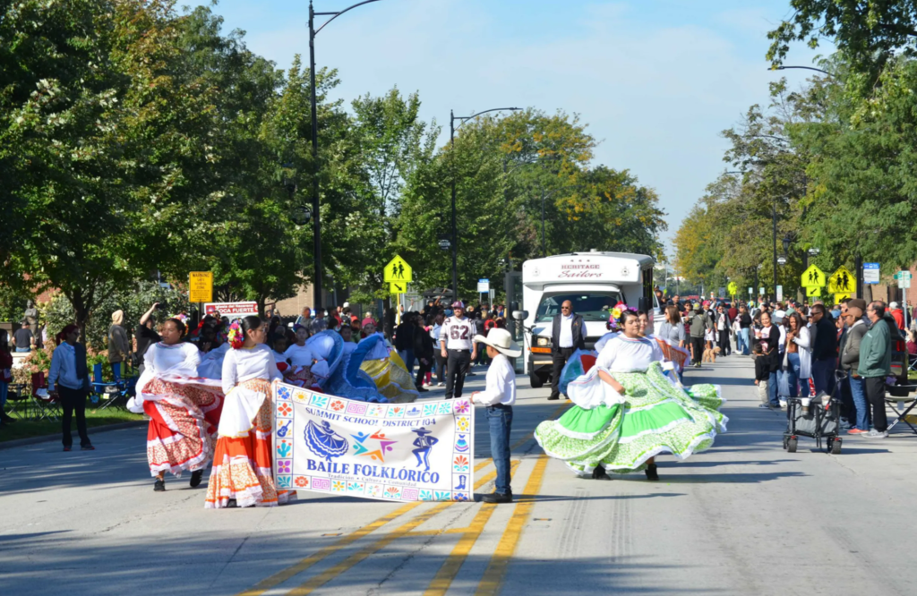 Colorfully dressed folklórico dancers carry a festival banner and twirl their skirts on a tree-lined street, while a bus and crowd look on in a festive mood.