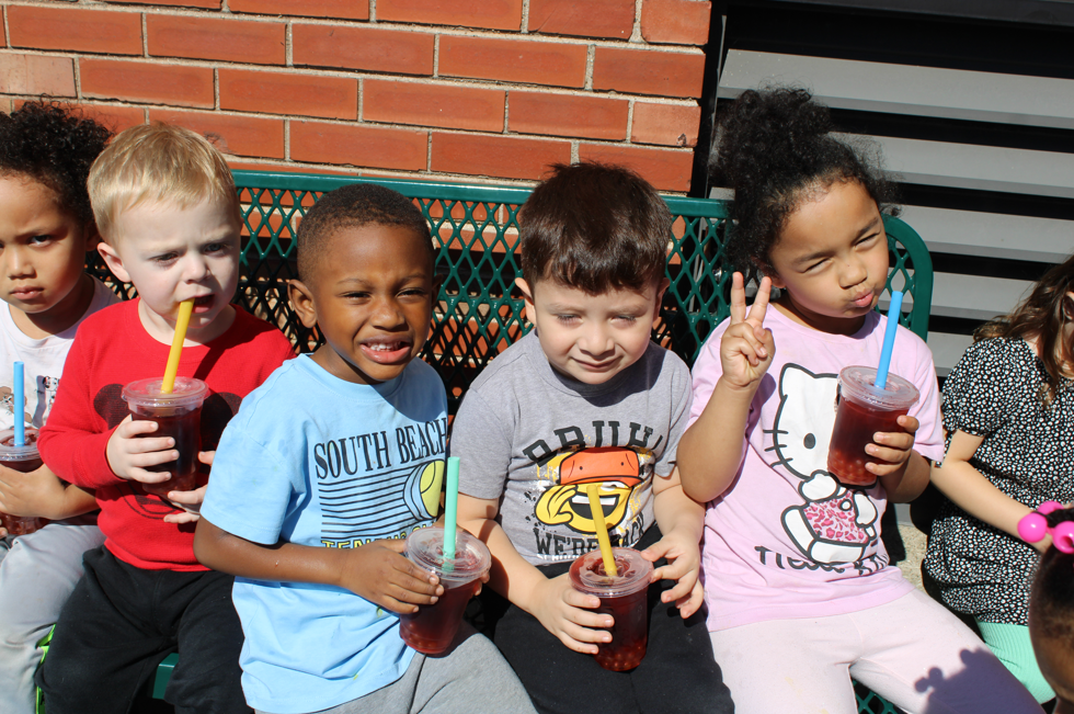 Six young children sit on a green bench outdoors, holding red drinks with straws; one flashes a peace sign against a brick wall in bright sunshine.