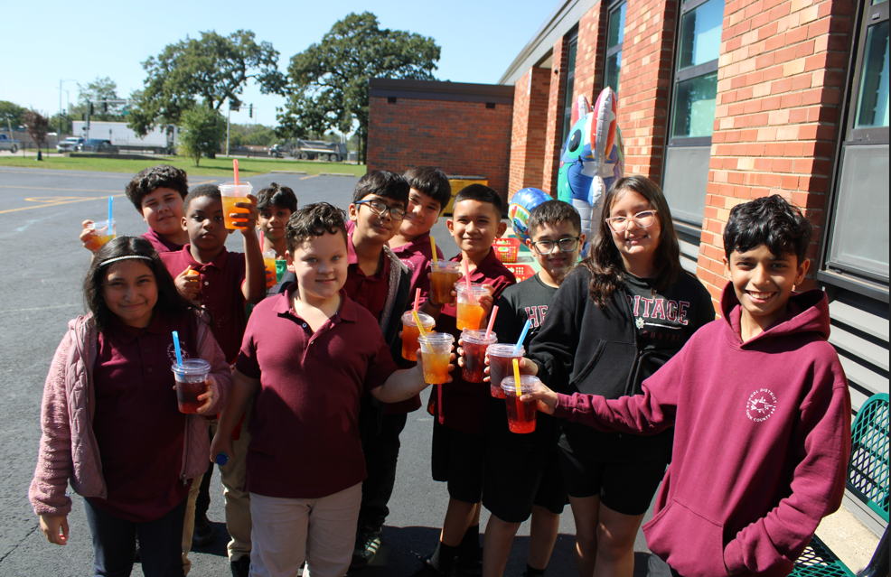 A cheerful group of students pose outside a brick school, holding colorful drinks; bright sunshine and smiles create a lively, celebratory mood.