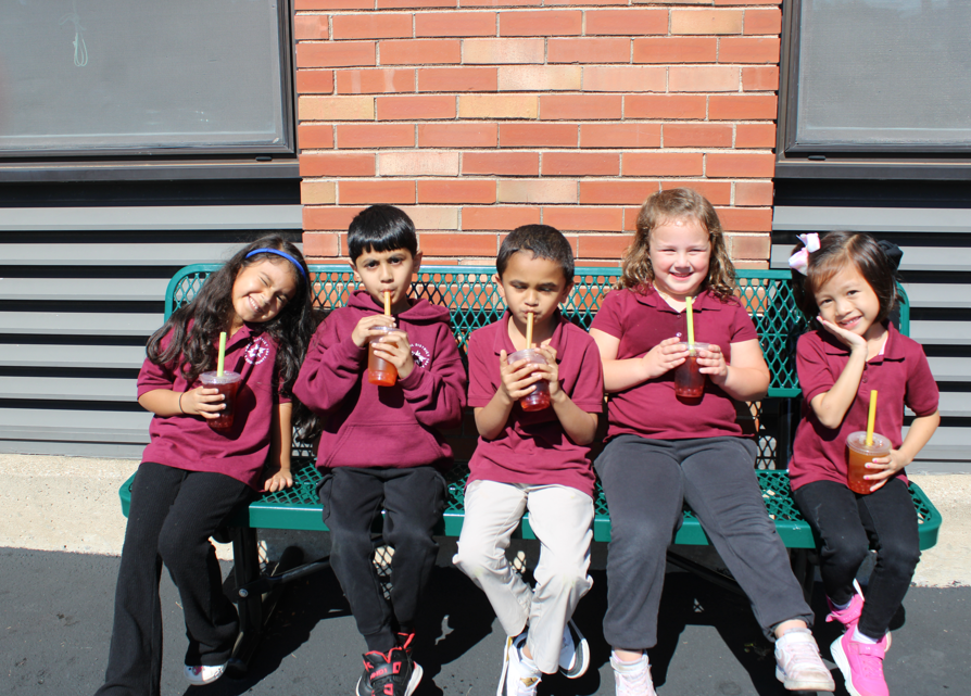 Five children sit on a green bench outside a brick building, sipping drinks in the sun; their smiles create a cheerful, playful mood.