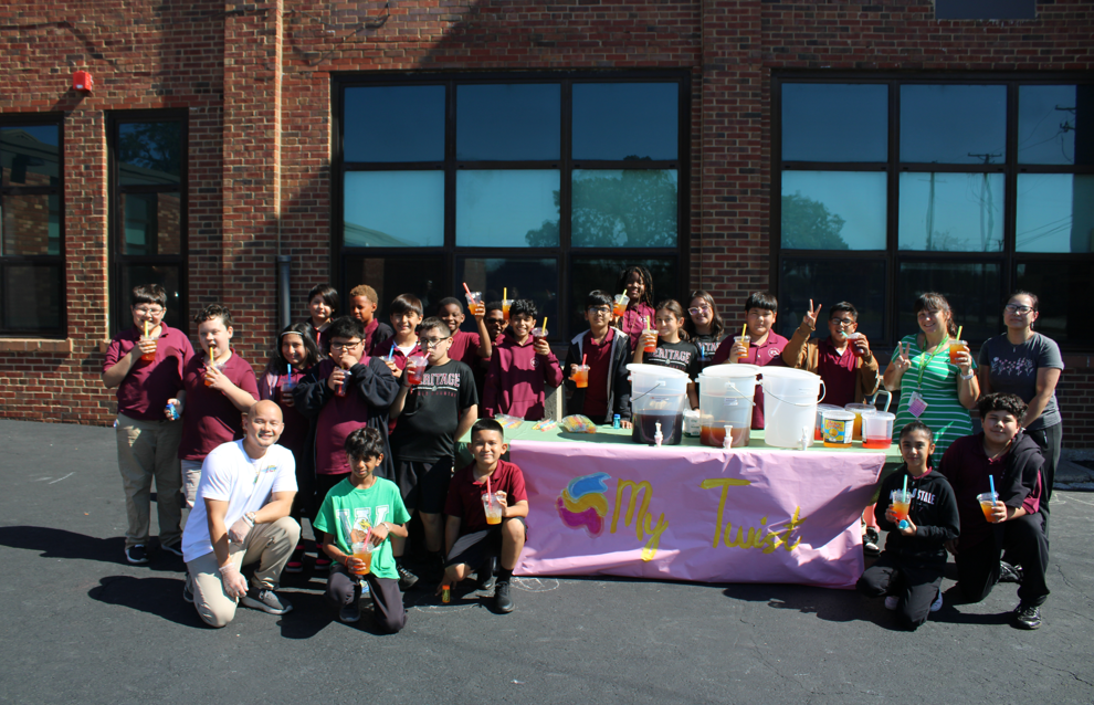 A cheerful group of children and adults gathers around a pink lemonade stand outside a brick building, with colorful drinks and handmade decorations.