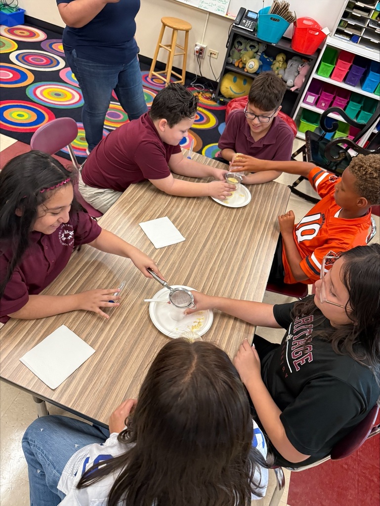 Students gather around a classroom table, using spoons and a strainer over small plates in a hands-on activity, with focused teamwork and colorful supply shelves behind them.