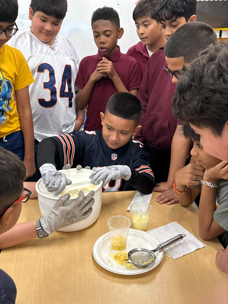 Students gather around a classroom science experiment as a boy in gloves handles a container, with cups and a strainer of yellow liquid nearby.