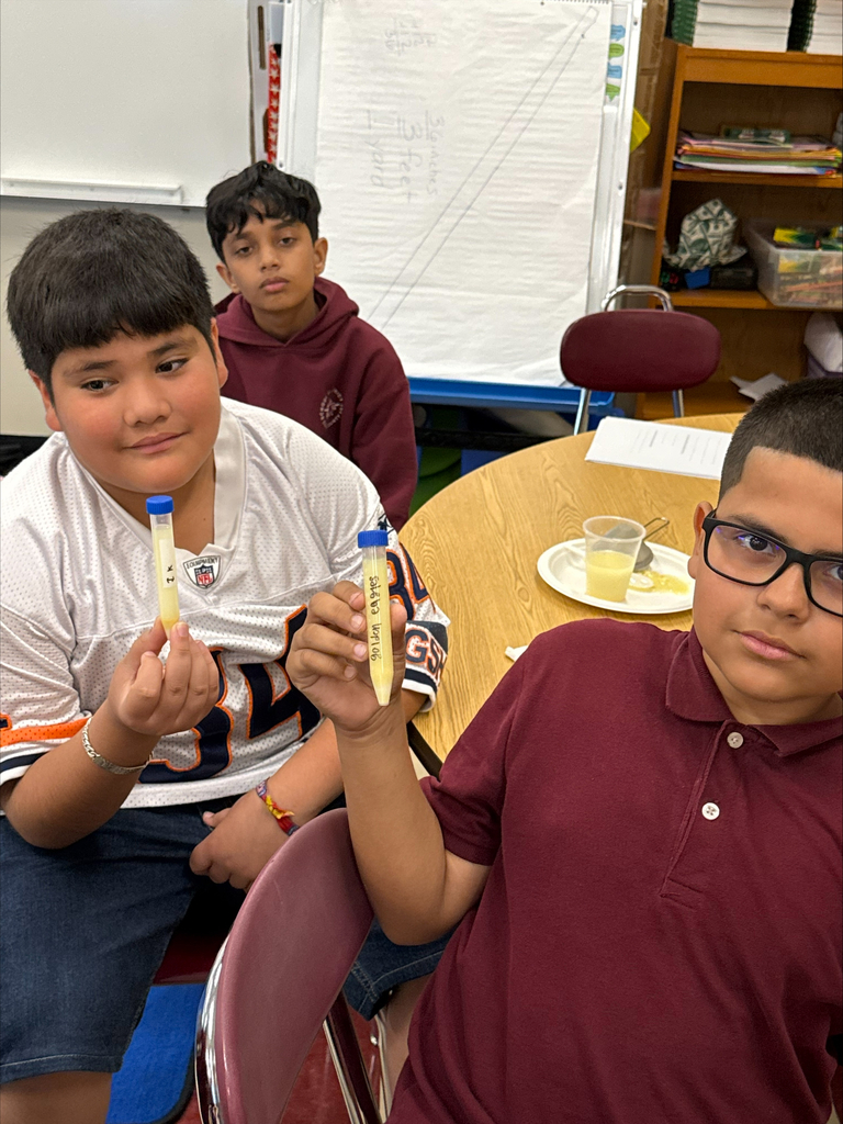 Three schoolboys sit in a classroom; two hold small tubes of yellow material beside a round table, with a whiteboard and shelves behind them.