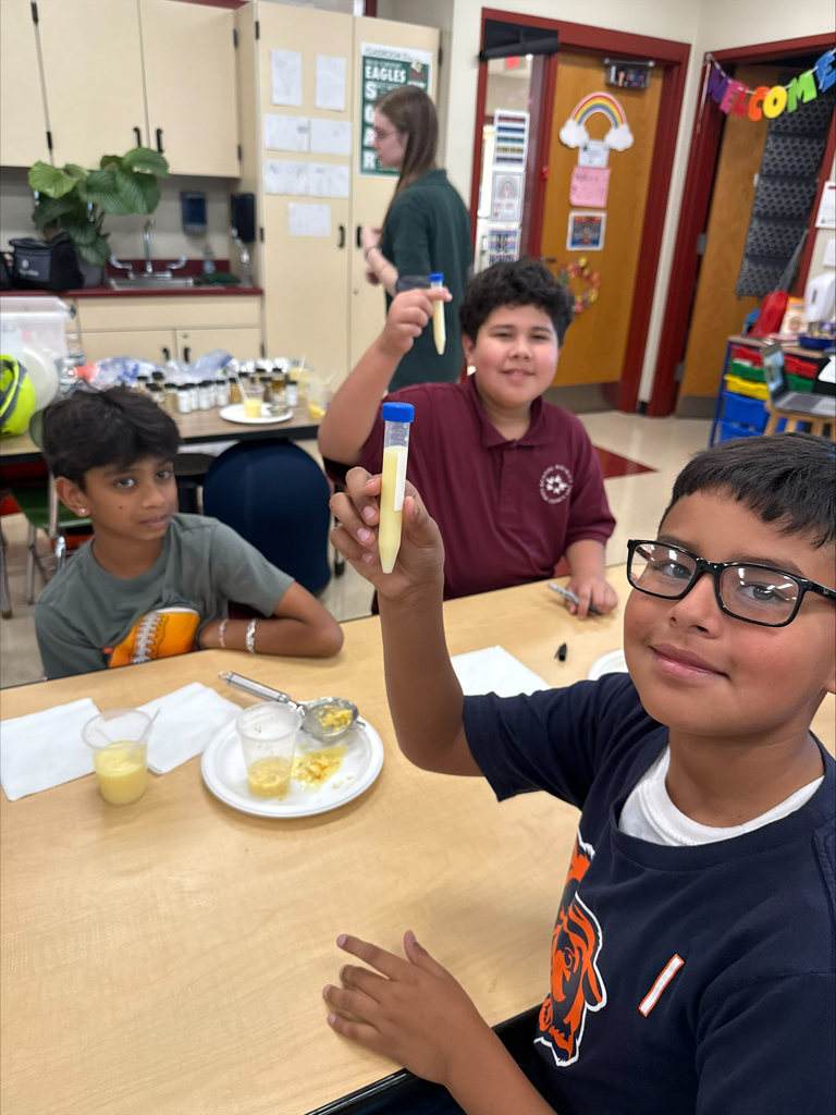 Three students gather at a classroom table for a hands-on experiment; a smiling boy in glasses holds up a test tube as classmates watch.