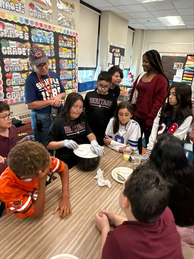 Students and teachers gather around a classroom table for a hands-on food activity, with one student wearing gloves and holding a plate; colorful posters fill the background.