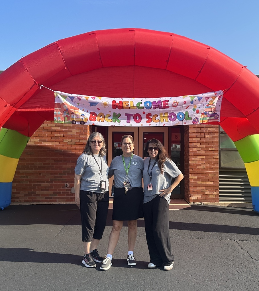 Three smiling school staff members pose beneath a bright red inflatable rainbow arch and “Welcome Back to School” banner outside a brick school on a sunny day.