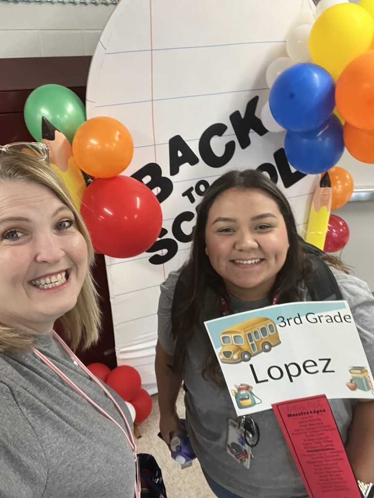 Two smiling women pose at a Back to School display with colorful balloons; one holds a “3rd Grade Lopez” sign, celebrating the new school year.