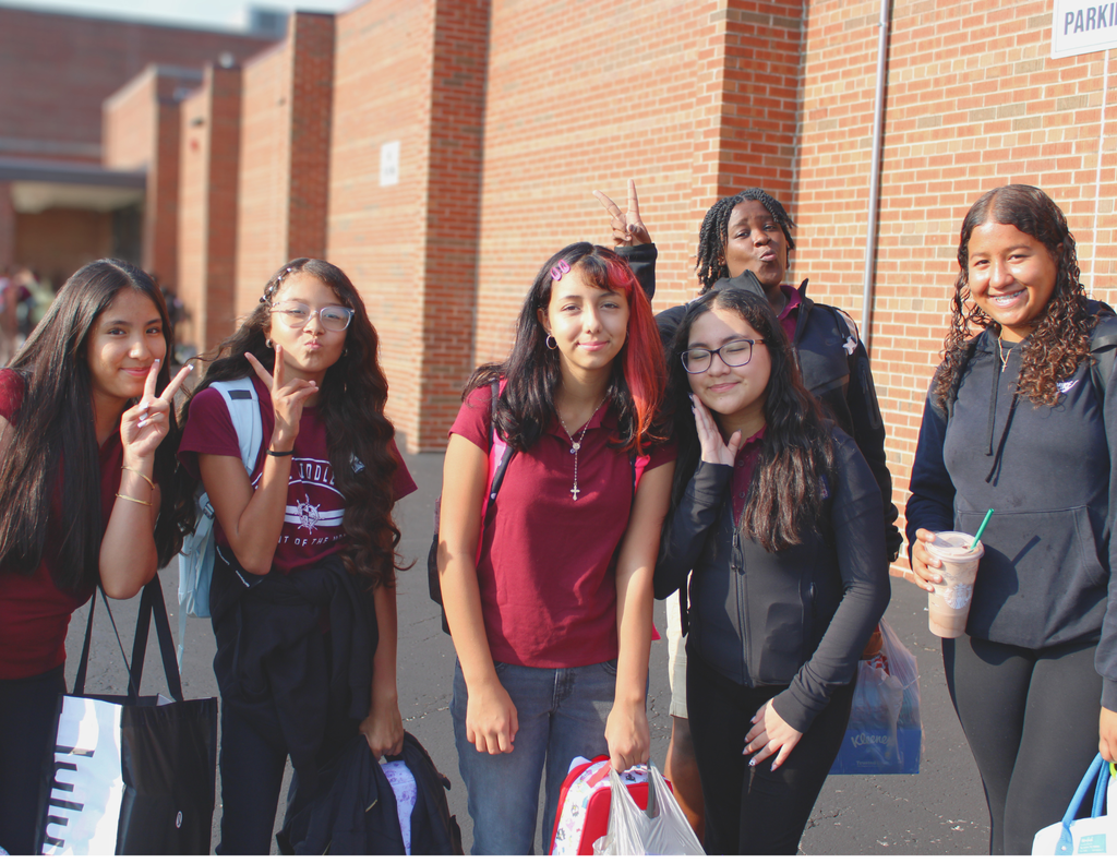 Six smiling students pose outside a brick school, flashing peace signs and holding backpacks and drinks in bright sunlight, creating a cheerful, friendly mood.