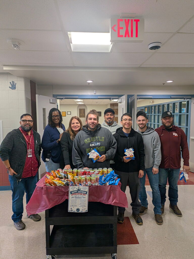 Seven smiling staff members pose behind a table stocked with snacks and drinks in a school hallway, creating a warm, community-focused atmosphere.