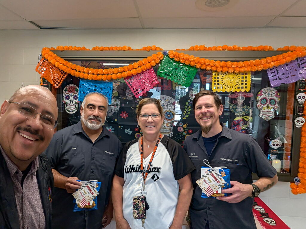 Four smiling coworkers pose before a vibrant Día de los Muertos display with orange marigold garlands, papel picado, and colorful skulls, conveying a festive mood.