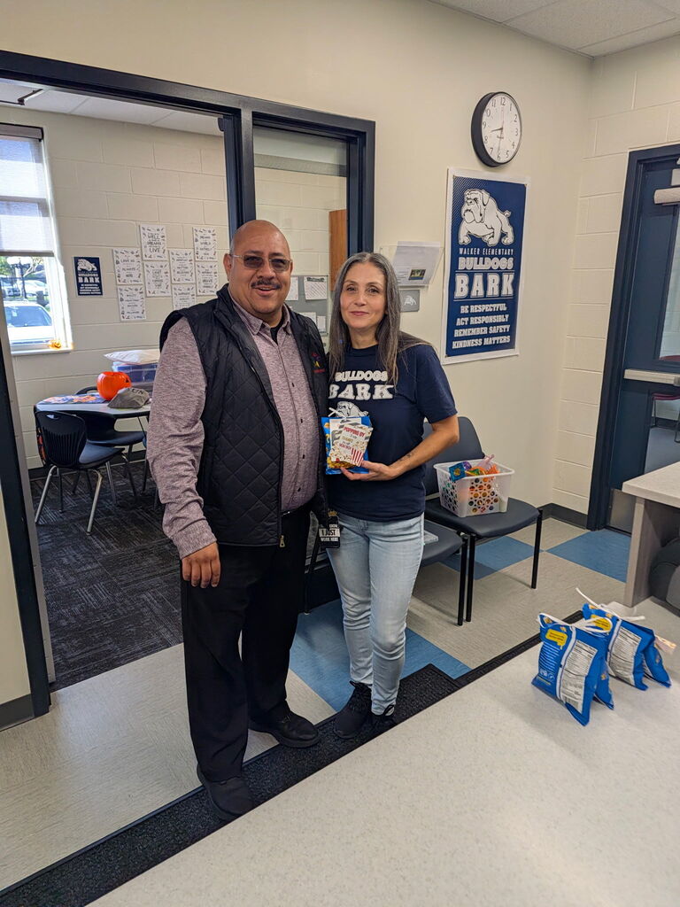 Two smiling adults stand in a school hallway, holding a snack bag beside a Bulldog Bark sign and more donated snacks, conveying a warm, generous community spirit.