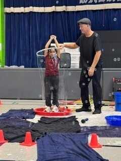 A man helps a child make a giant soap bubble with a large circular frame during a playful, hands-on science activity in a school gym.
