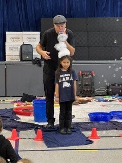 A man in a cap steadies a fluffy white dog above a child on a mat, surrounded by cones and training gear in an indoor space.