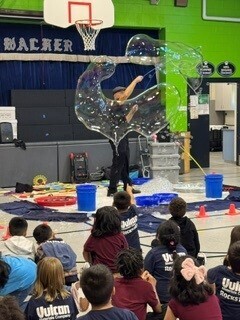 In a school gym, students watch a performer suspended inside a shimmering, transparent bubble, creating a magical, wonder-filled spectacle.