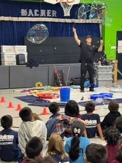 A science-show presenter gestures beneath shimmering giant soap bubbles onstage as children watch, creating an energetic, wonder-filled mood.