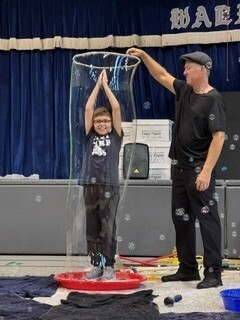 A smiling child stands inside a tall transparent bubble on a stage as an instructor reaches over to guide it; blue curtains and scattered bubbles set a playful mood.