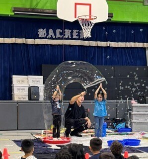 A martial arts instructor and two children practice staff techniques beneath a basketball hoop in a gym, surrounded by equipment; the scene feels lively and focused.