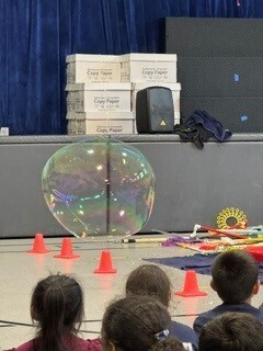 Children watch a large, shimmering soap bubble during an indoor science demonstration, with orange cones and experiment supplies on the floor.