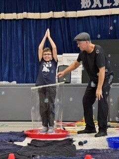 A smiling boy stands on a red plastic disk with hands pressed together overhead as a man steadies a tube, during a playful school science demonstration.