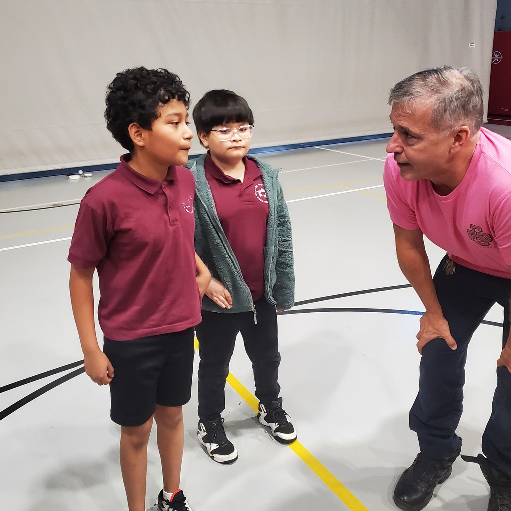 Two schoolchildren in maroon uniforms talk with a coach in a pink shirt, who crouches to their eye level in a gym, creating a warm, attentive mood.