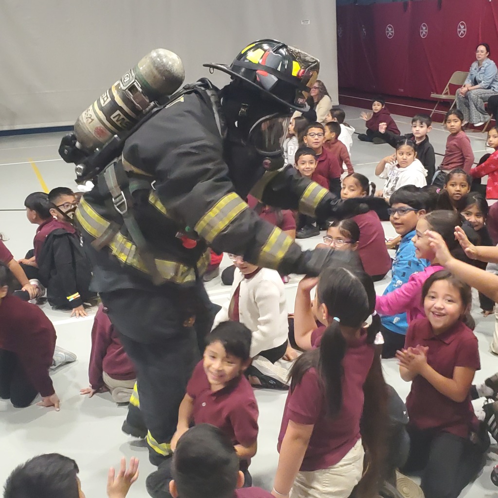 A firefighter in full protective gear kneels among seated schoolchildren, sharing a safety demonstration; smiling, attentive faces create an excited, welcoming mood.