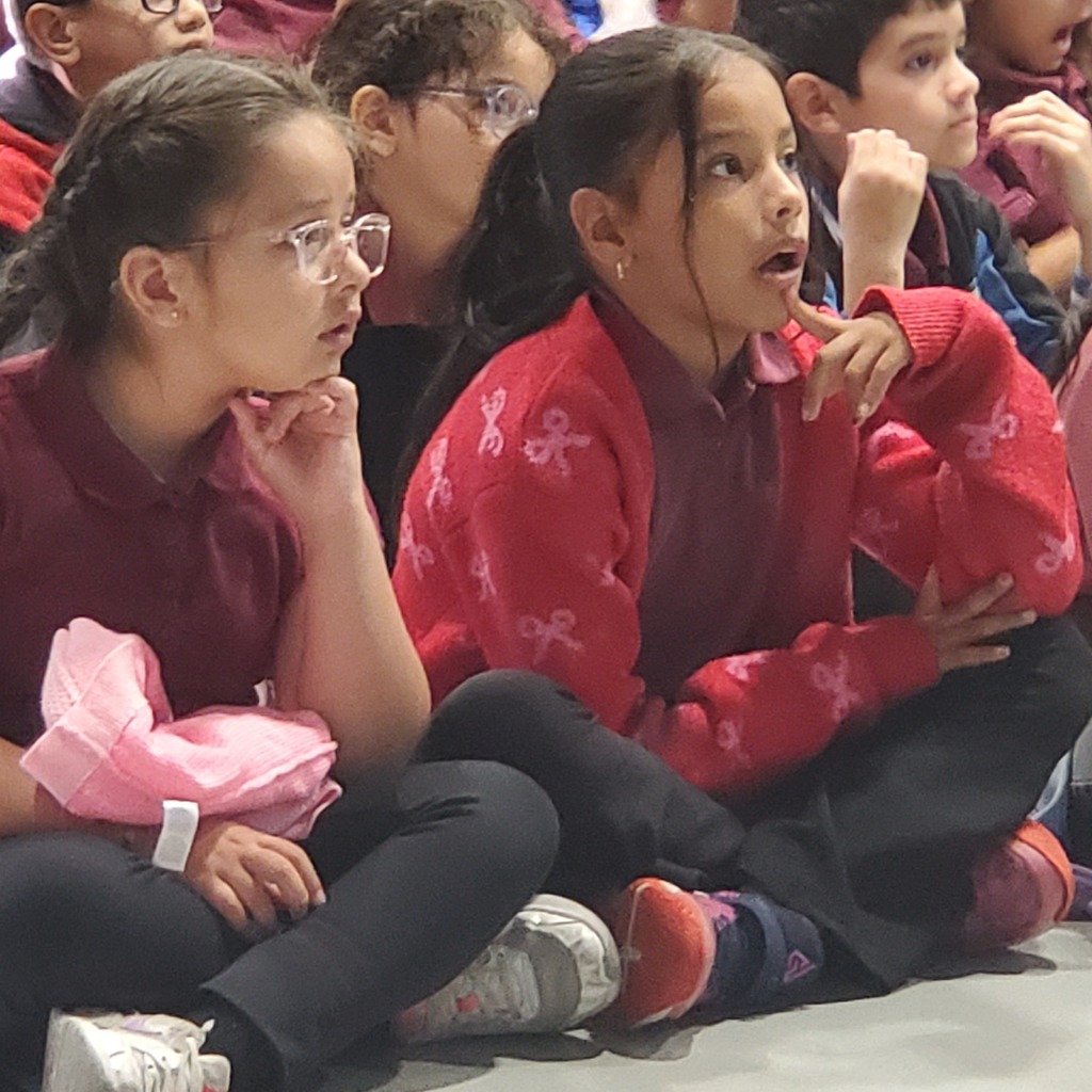 Children sit closely together at an indoor gathering, watching something off-camera with focused, curious expressions; one girl in a red sweater looks surprised.