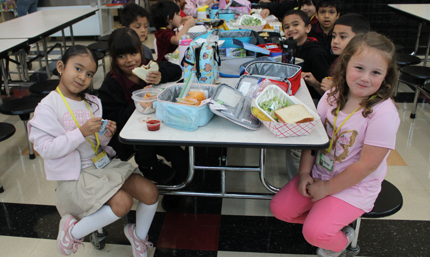 Elementary school students sit around a cafeteria table with lunch bags, sandwiches, fruit, and snacks, smiling at the camera in a cheerful, friendly mood.