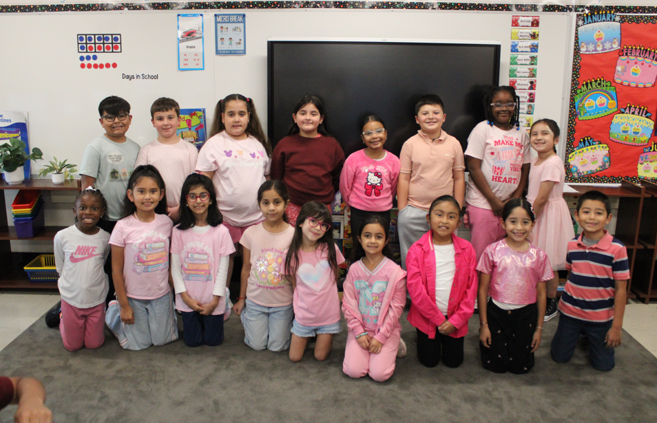 A diverse group of smiling schoolchildren pose in two rows before a classroom screen, many wearing pink, creating a warm, cheerful group portrait.